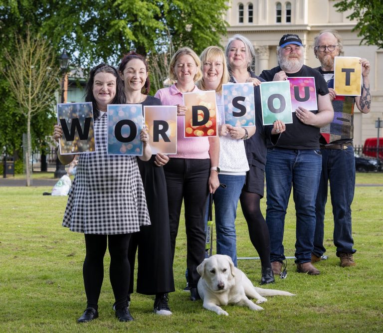 Seven people stand at a diagonal each holding a letter which together spells out the word "Words Out". They are standing in the middle of a small green.