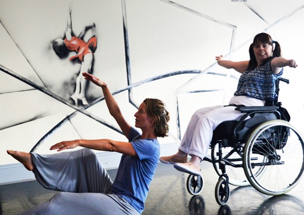 Picture of two female dancers, one of which is a wheelchair user with her arms stretched wide. The other dancer is sitting on the floor with her arms stretched in front of her.