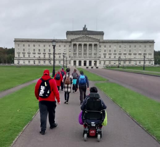 A picture of a group of people one of which is a wheelchair user walking along the footpath towards Stormont Parliament Buildings.