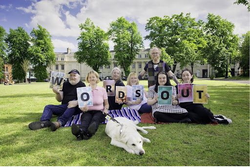Six Open Arts participants sit on a green surrounded by trees. They each are holding a letter, which spells "words out." Debbie the guide dog is lying in front of everyone.