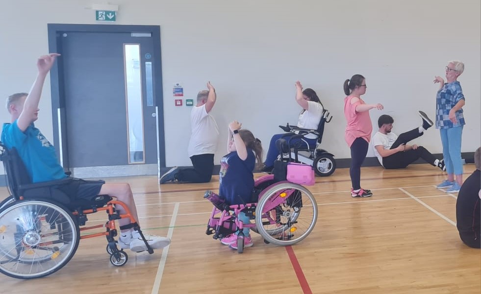 Picture of 6 teenagers from Mae Murray, three of which are wheelchair users are taking part in an intensive dance workshop. There are three wheelchair users and three non wheelchair users. All have their arms in the air and are moving animately.
