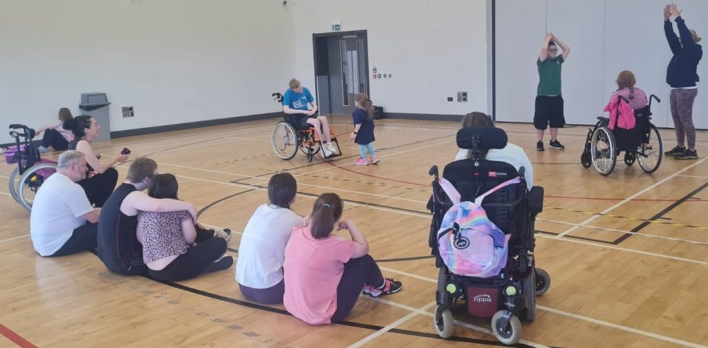 Six teenagers are sitting with their backs to the camera in a semi-circle watching four of their peers in the middle dancing in pairs. Two of the teenagers are wheelchair dancers.