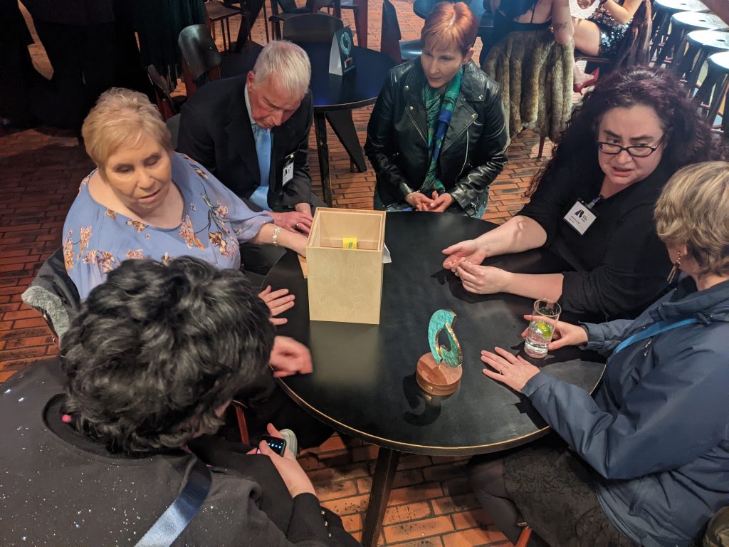 Margaret is sitting at a round table with her award. It is very tactile and is constructed of two crescent shapes intertwined mounted on a round wooden base. Also at the table chatting are members of Open Arts board and staff.
