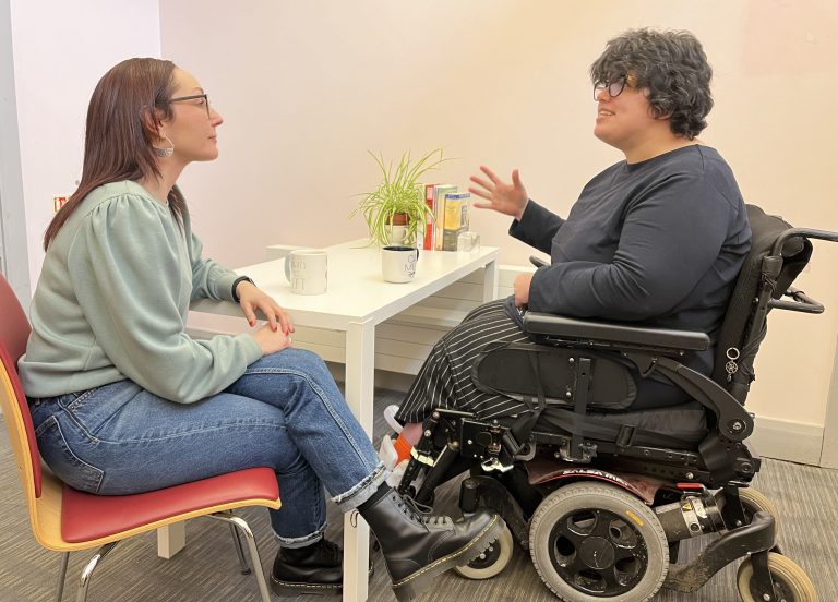 Jemma & Cinzia are sat a table engaging in conversation. Jemma has long dark hair and is wearing jeans and a green jumper. Cinzia has short dark hair and is wearing a black top and stripy trousers, she is a powerchair user.