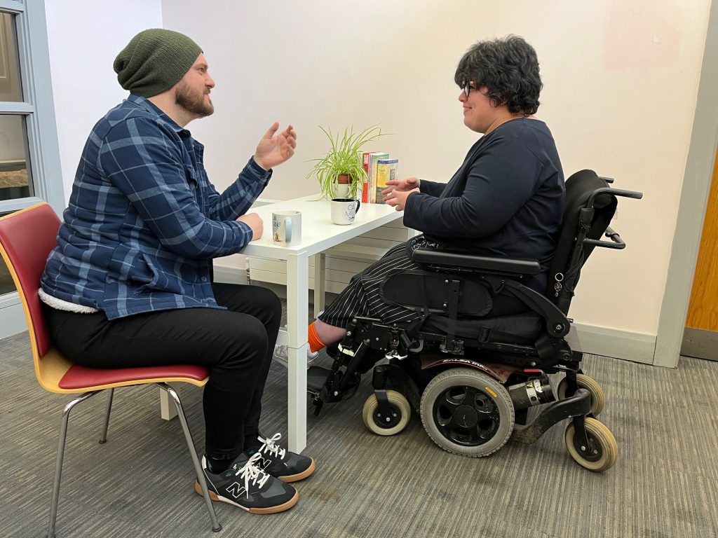 Ciaran & Cinzia are facing each other at a table and engaging in a conversation. Cinzia is a powerchair user with short dark hair and is wearing a black top and stripy trousers. Ciaran is wearing a moss green beanie hat and is wearing a blue checked shirt and black jeans.