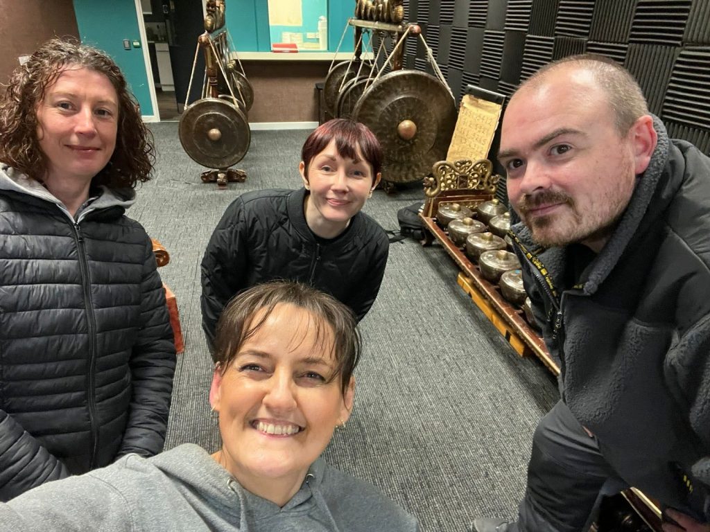 A selfie of Eileen, Bev, Jemma and Andy standing in the new unit in Connswater Shopping Centre where the Gamelan is now housed. Behind them you can see 2 large racks with Gongs hanging from them.