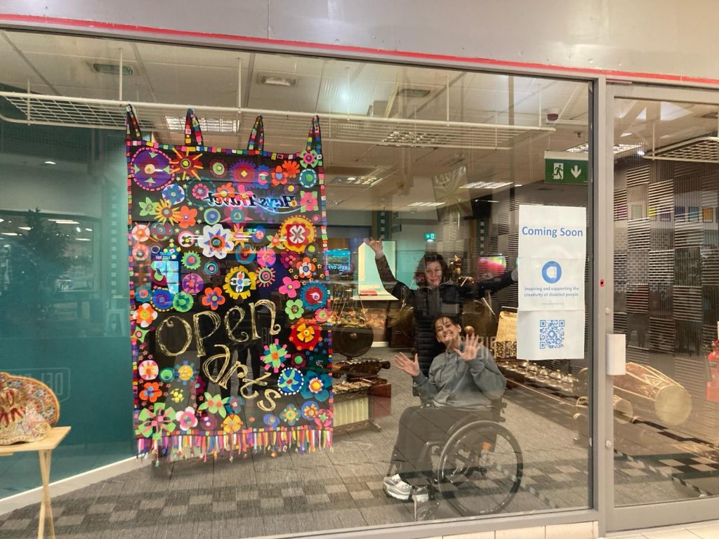 Eileen and Bev are waving enthusiastically and happily in the window of the new Connswater Shopping Centre Unit. There is a black banner hanging in the window that has bright and colourful flowers and the words "Open Arts" written in gold. There is a sign saying beside the door coming soon.