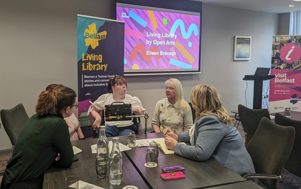Mary Lou is with her sister and is using her assistive communication device to chat with two women attending the Living Library event. Behind them are two penguin stands with promotional information about Living Libraries and Visit Belfast.