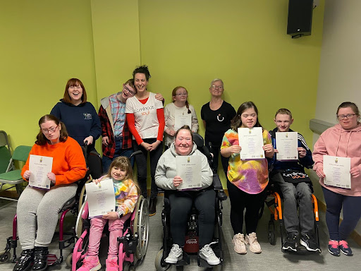 Nine young people from Mae Murray's Teen Hub are holding their Arts Award Certificates with Luminous Soul dancers Helen & Linda. They are all smiling and laughing at the camera.