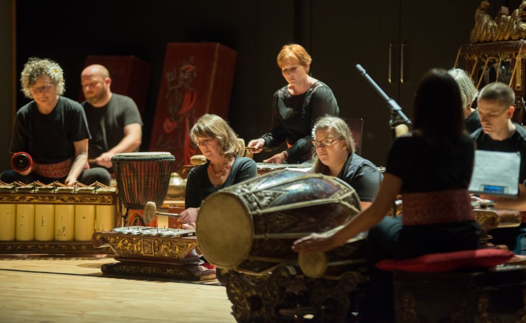 Seven gamelan members play instruments on stage at Julan Julan production whilest being conducted.