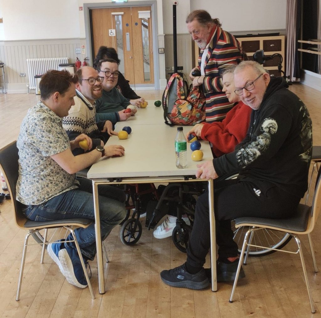 Seven participants are sitting at either sides of a long rectangular table. They have two multi-coloured juggling balls each in front of them.