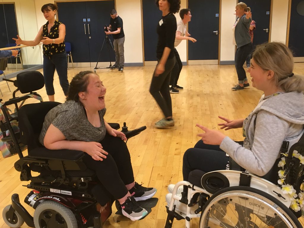 A group of young people are taking part in a dance workshop. In pairs they are mirroring what the other person is doing. There is a lot of laugher and smiles.