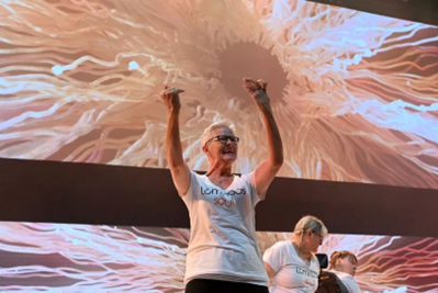 Picture of Linda Fearon performing at the One Young World opening ceremony. She is wearing a white t-shirt with the Luminous Soul logo and her arms are stretched upwards. She is smiling.