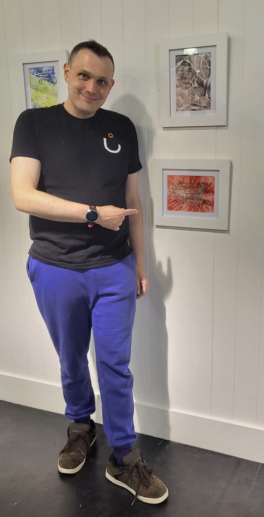 Darren our participant is wearing blue jogging bottoms and a black t-shirt. He is pointing to a piece of artwork that he has created and is hung on the wall. The picture is small in size and is of burnt orange, red and white colours