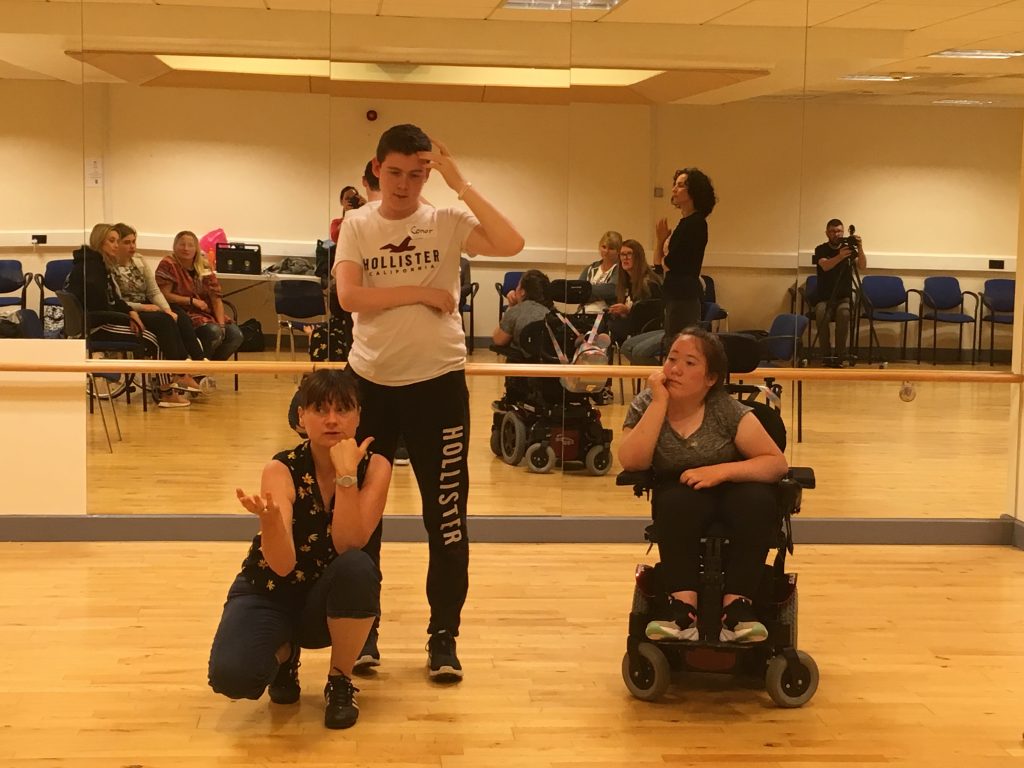 Two young people, one who is a wheelchair user are in front of an audience that you can see reflected in mirrors behind them. They are also accompanied by Orla the facilitator. She is kneeling down with her head resting on her fist.