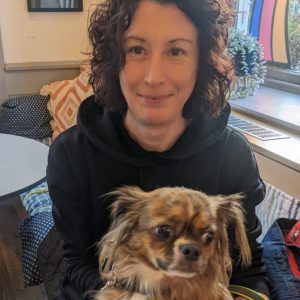 Open Arts CEO Eileen is sitting in a cafe holding her brother's small dog Teddy on her lap. He has longish tan and back hair with a white fluffy belly.