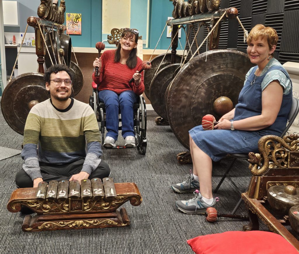 Three musicians from the Open Arts gamelan group sit on chairs nestled between two racks filled with suspended various sizes of metal gongs. They are holding wooden beaters and facing forward.