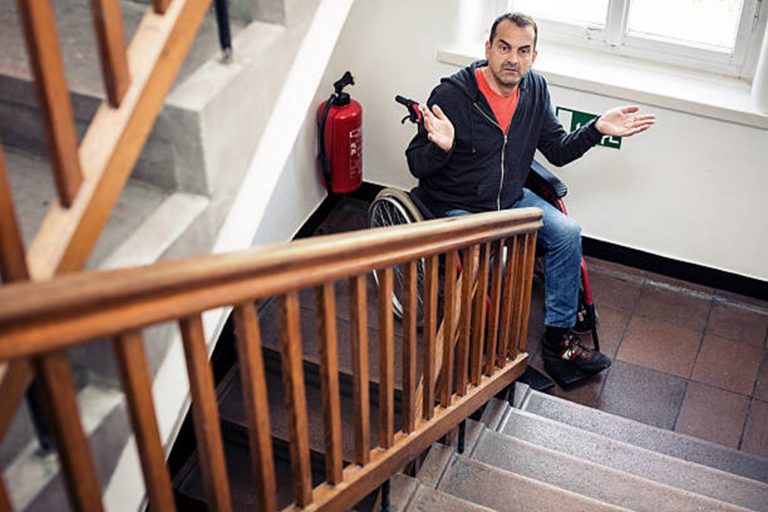 Man sitting in a wheelchair looking up at stairs above him. He is putting his hands out to his sides in frustration.