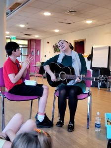 Musician Bronagh Broderick is sitting on a chair with her guitar on her lap. Beside her is a young boy engaging in her singing.
