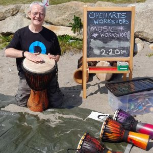 Musician Jonny McGeown is on a beach. There is a small chalkboard beside him that says "Open Arts workshop music 2:20pm." He is knelt down with a djembe drum in front of him. There are three smaller ones sitting on the sand in front of him.