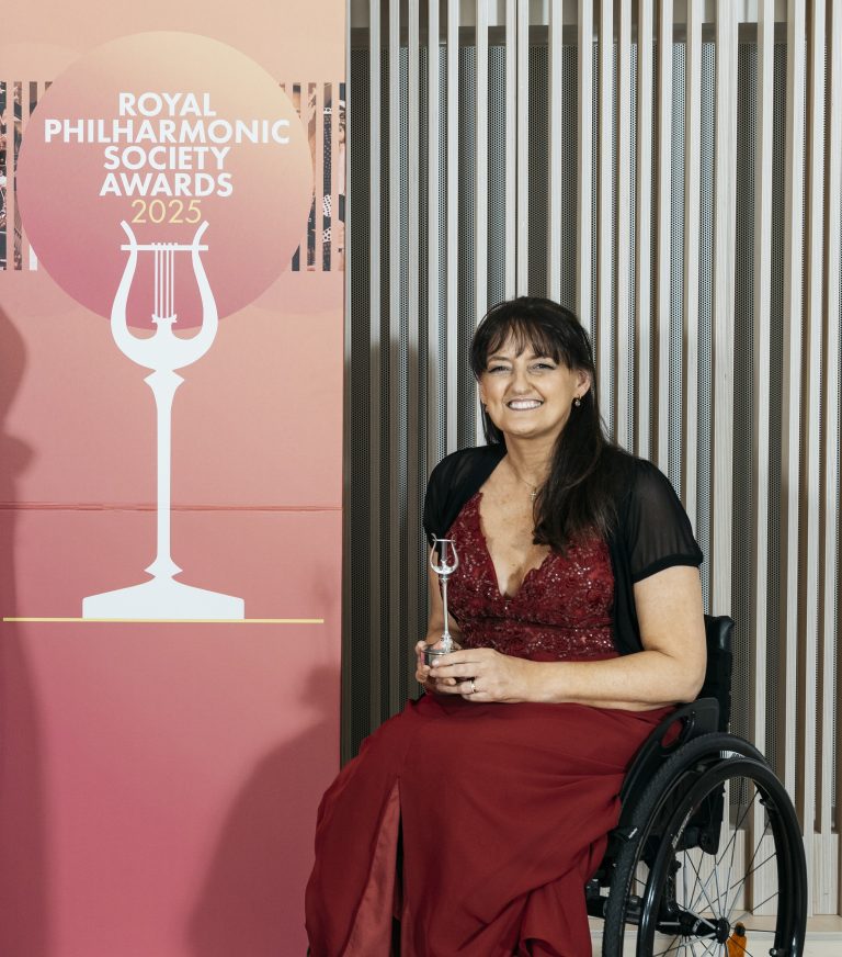 Photo of Bev, sitting in her wheelchair in a long red formal gown, it has red beaded details on the bodice. She is holding a silver award in the shape of a harp, smiling at the camera.