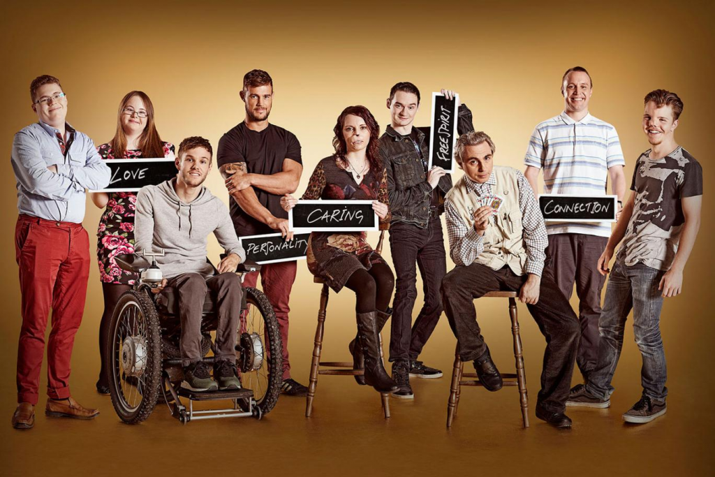 ALT TEXT: 9 individuals are smiling at the camera, they have different visible and non-visable disabilities ranging from using a wheelchair, to having a facial difference. They are holding signs that say “love, personality, caring, freespirit, connection.”