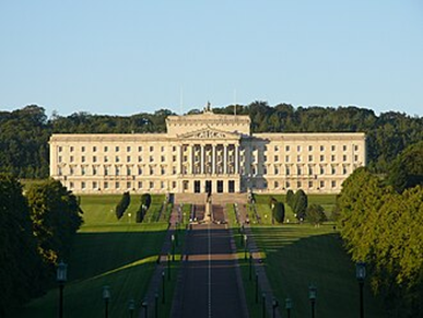 Stormont Parliament Buildings. A long, white, rectangular building with ornate pillars and décor on the outside. It is photographed in the sun, with trees surrounding it and a tarmac walkway up to the building.