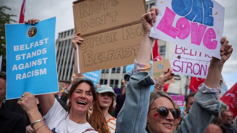Photo of people at protest holding signs that say “Belfast stands against racism”, “Riots don’t speak for Belfast”, and “One love.” They are smiling and shouting in front of Belfast City Hall.