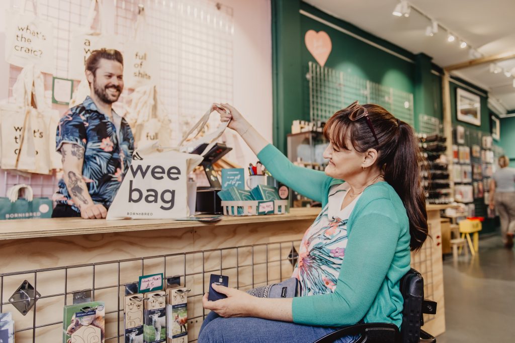 A female wheelchair user at a shop counter holding a beige tote bag that reads “wee bag – BORN&BRED.” She is smiling at the shop assistant.