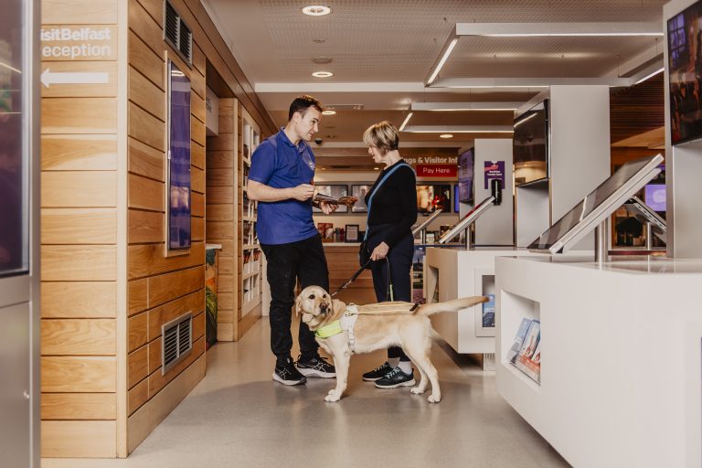 A guide dog user speaks with a staff member at a modern information desk. The space has wooden panelling, white counters with digital screens and signage for Visit Belfast reception and visitor bookings.