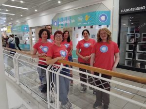 Six staff from the Open Arts Accessibility hub stand outside the shopping centre unit. They are all wearing pink t-shirts with the Open Arts logo.